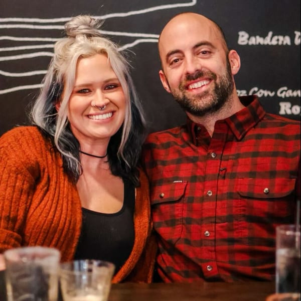 Smiling woman with blonde hair and man with beard in plaid shirt at a restaurant.