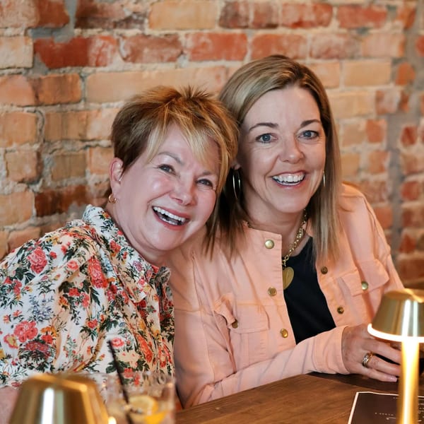 Two smiling women sitting at a table with drinks, against a brick wall.