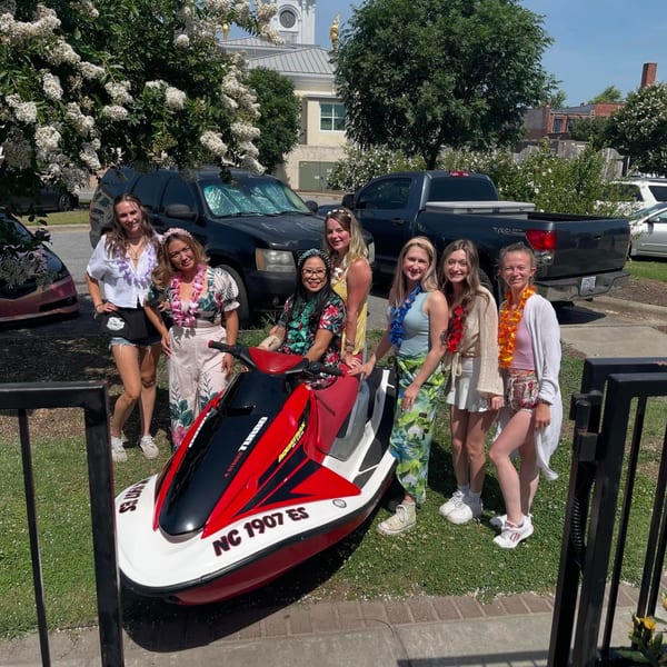 Seven women in summer attire and leis pose around a red and white jet ski on a sunny day.