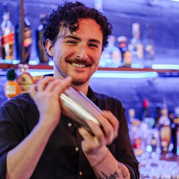 Smiling bartender with curly hair and mustache shaking a cocktail shaker in a dimly lit bar.
