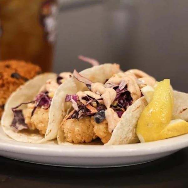 Two shrimp tacos with purple cabbage, sauce, and lemon wedges on a white plate.