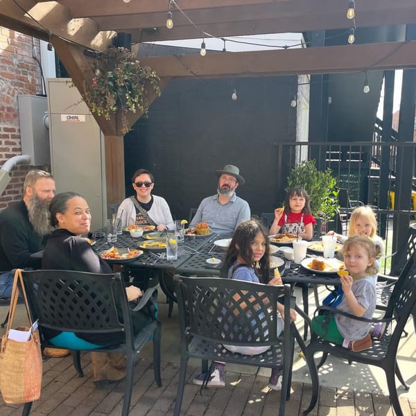 A family with children enjoys a meal together at an outdoor restaurant patio on a sunny day.