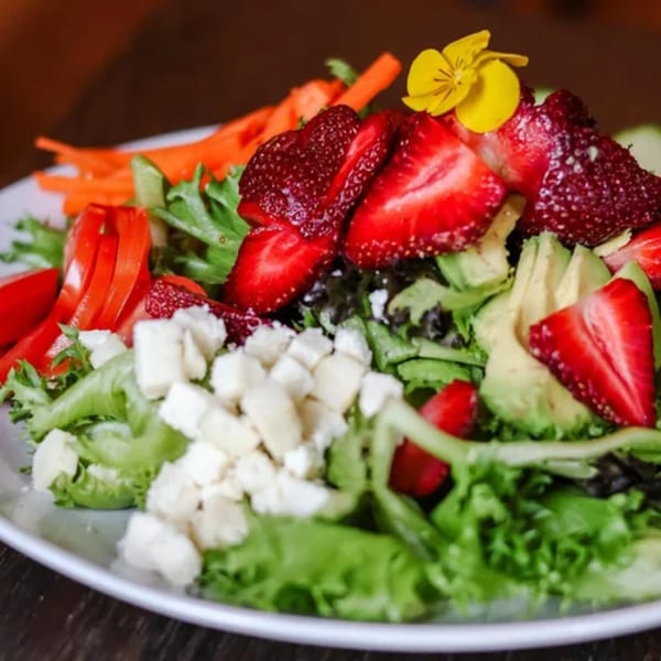 A vibrant salad with strawberries, avocado, feta, carrots, and red peppers, garnished with a yellow flower.