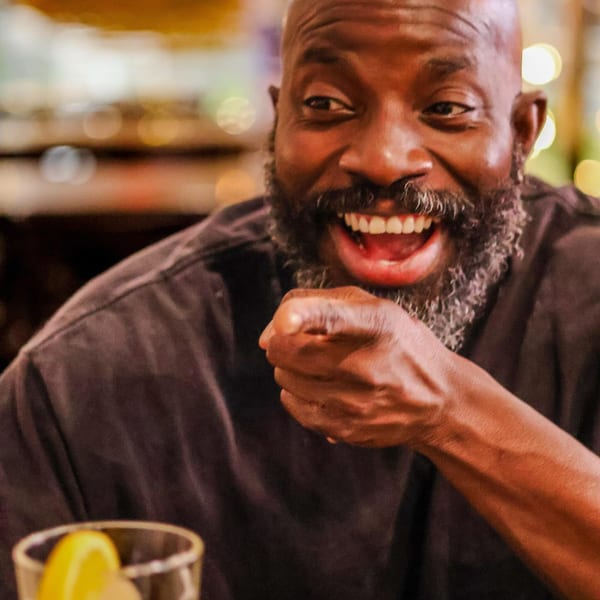 A man with a beard laughs and points, with a drink in the foreground.