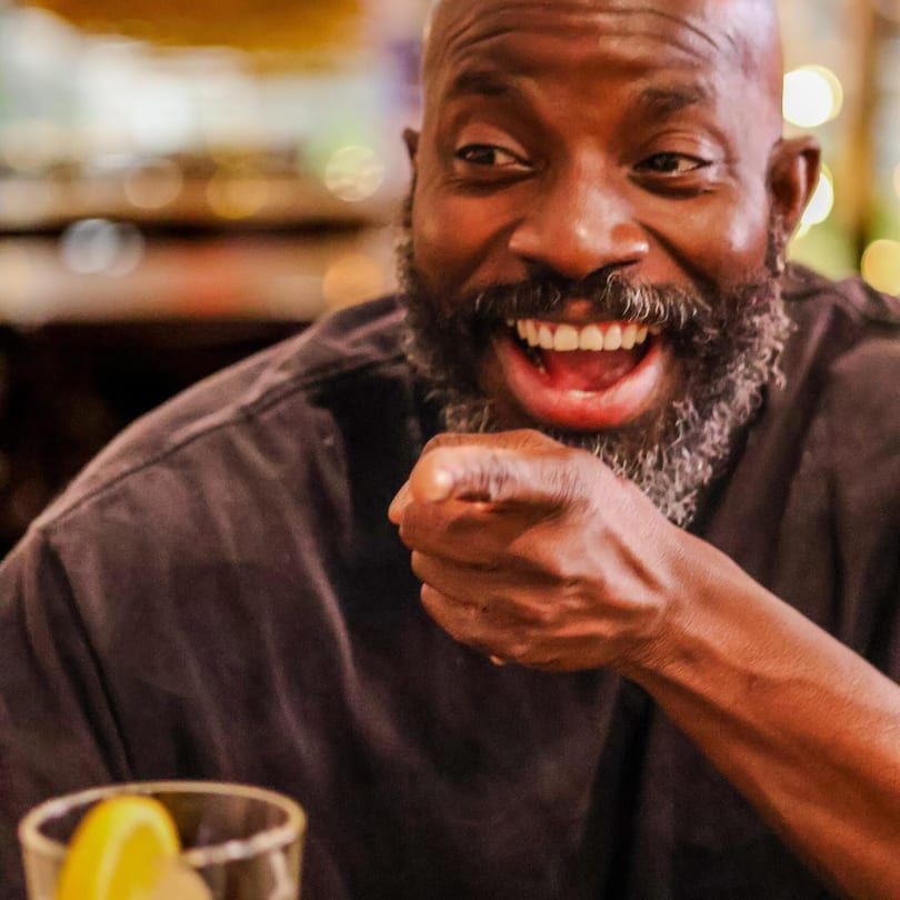 A man with a beard laughs and points, with a drink in the foreground.