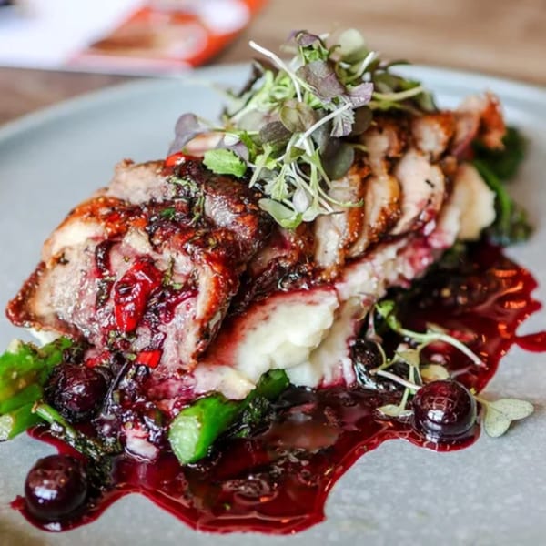Sliced duck breast with berry sauce, mashed potatoes, asparagus, and microgreens on a gray plate.