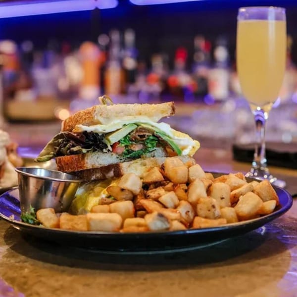 A breakfast sandwich with avocado, egg, and bacon, served with home fries and a mimosa.