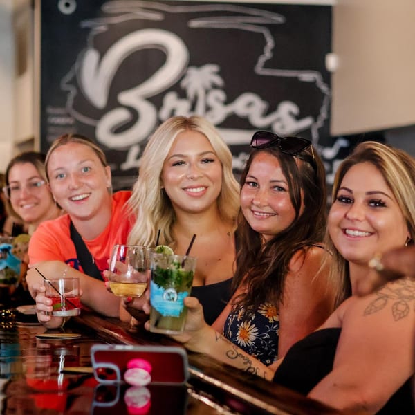 Five smiling women holding cocktails at a bar with a "Brisas" sign in the background.