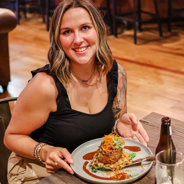 A smiling woman in a black top holds a plate with a steak dish topped with crispy onions.