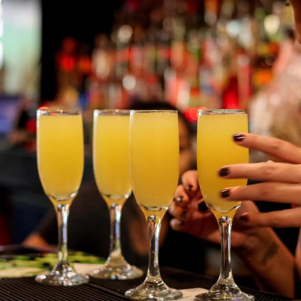 Four champagne flutes filled with mimosas on a bar, with a person's hand reaching for one.